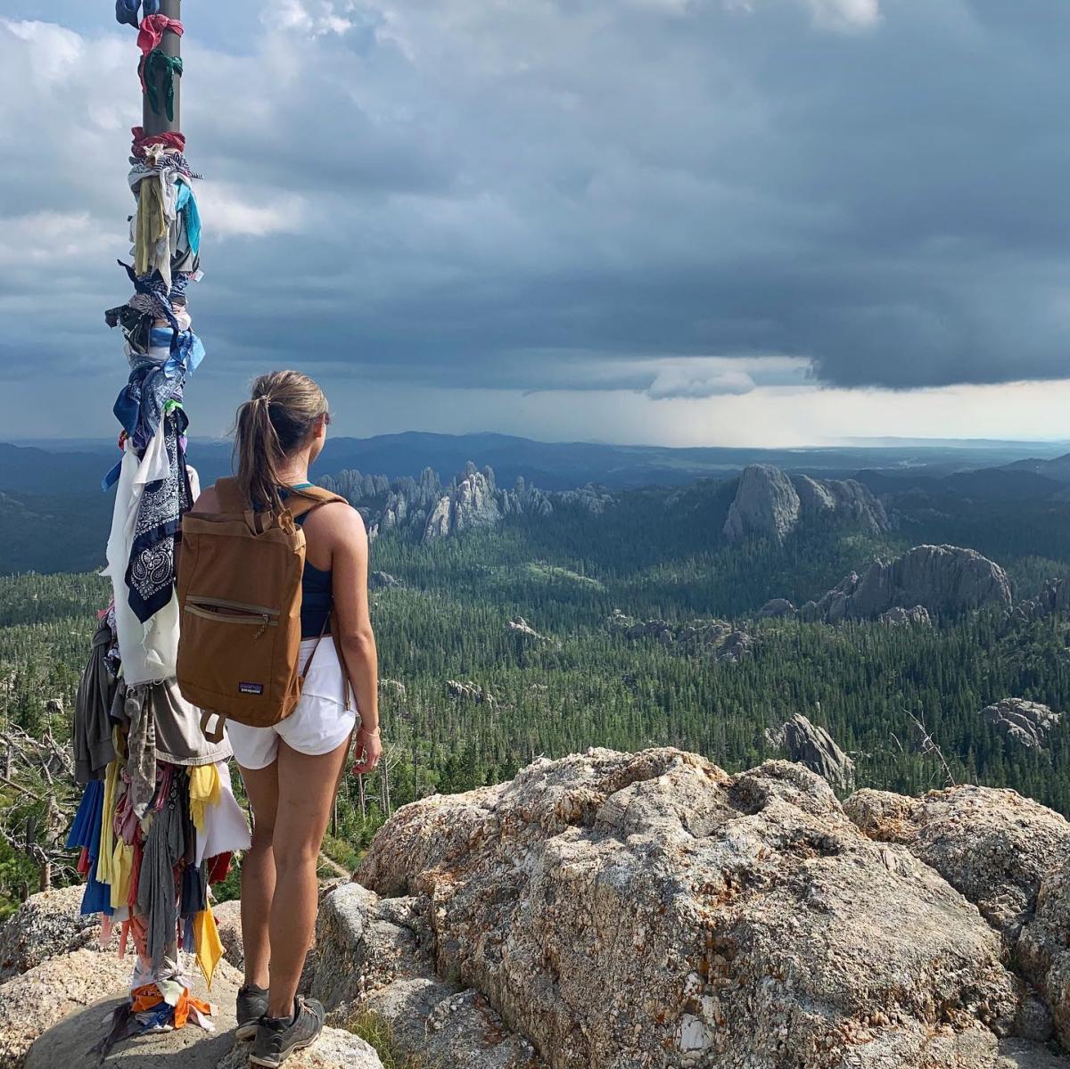 A woman with a brown backpack stands at the top of Black Elk Peak, gazing at a vast, forested landscape and distant mountains under a dramatic, cloudy sky.