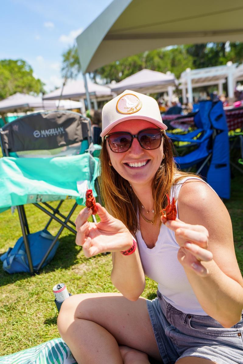 Woman seated on a blanket at the Mandeville lakefront smiles toward the camera while holding up two boiled crawfish, with festival tents and folding chairs behind her at the Crawfish Cook-off for a Cause.