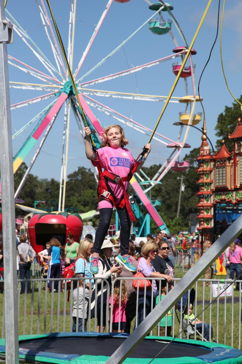 Young girl bounces on a bungee trampoline with a colorful Ferris wheel and midway rides behind her at the St. Tammany Parish Fair.