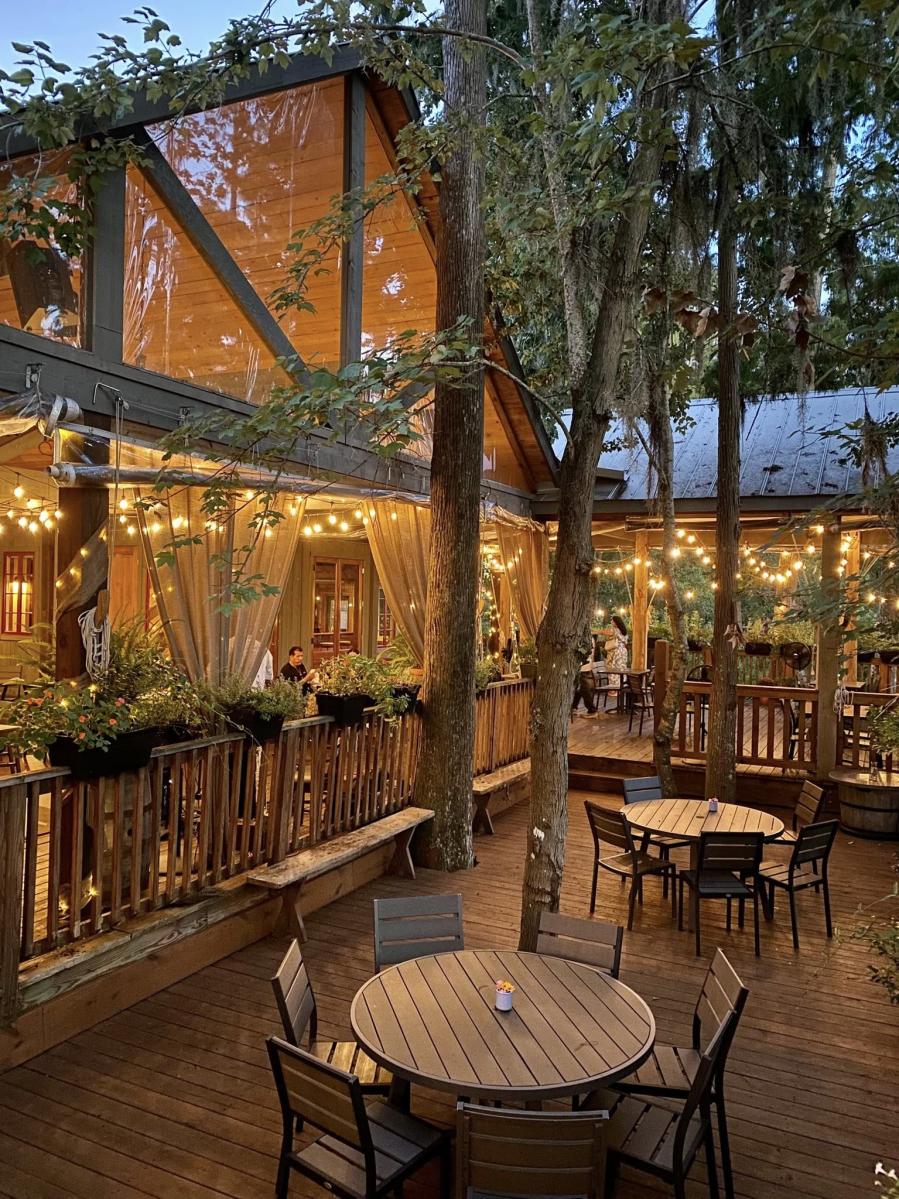 Two round, slatted wooden tables surrounded by matching wood and iron chairs, positioned on a sunken wooden deck with five tall trees rising through the floor. Behind is a raised, covered, wraparound deck lined with potted plants and gold string lights, attached to a building with large, reflective windows. It is twilight.