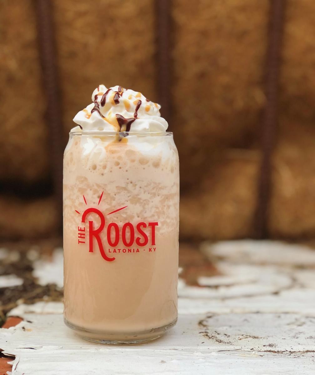 Image is of an iced coffee with whipped cream, caramel and chocolate syrup in a glass mug that says "The Roost, Latonia-KY".