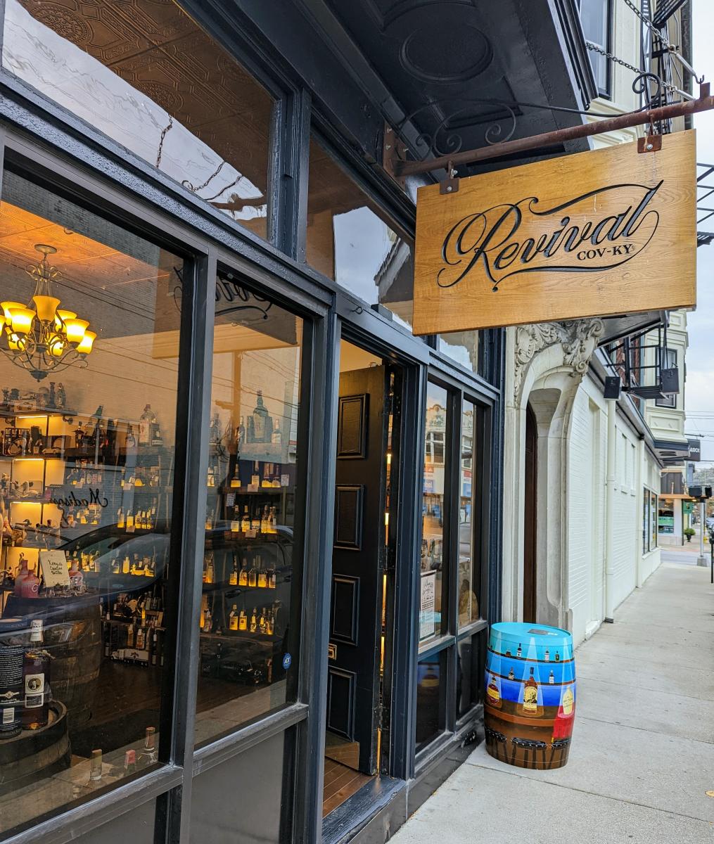 The glass windowed front of Revival Vintage Spirits with bourbon bottles visible through the windows and a wood sign with Revival written on it hanging over the door