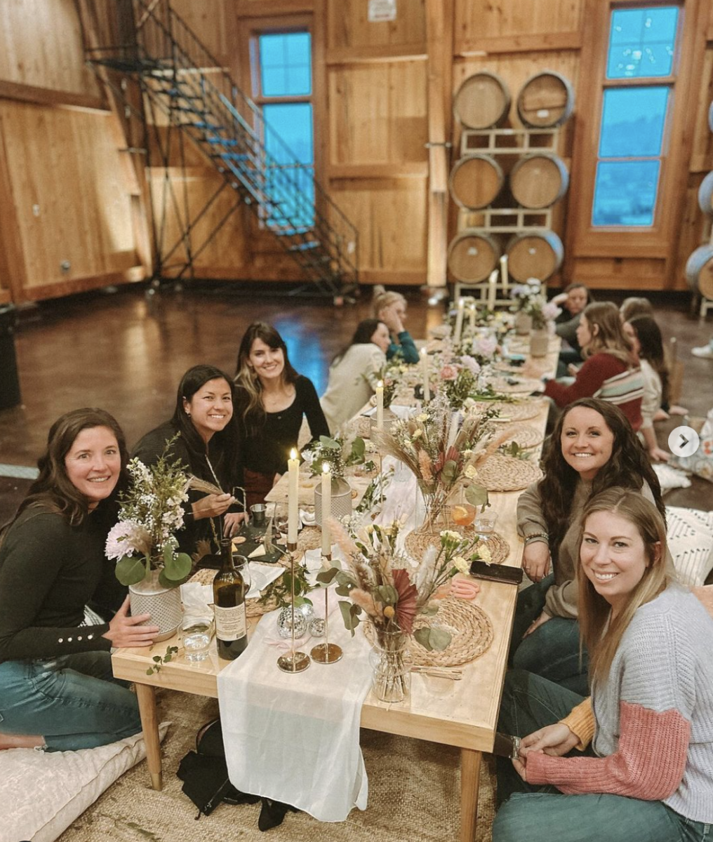 A group of women enjoying a picnic inside, sitting on the floor with a beautifully decorated picnic table with flowers and charger plates.