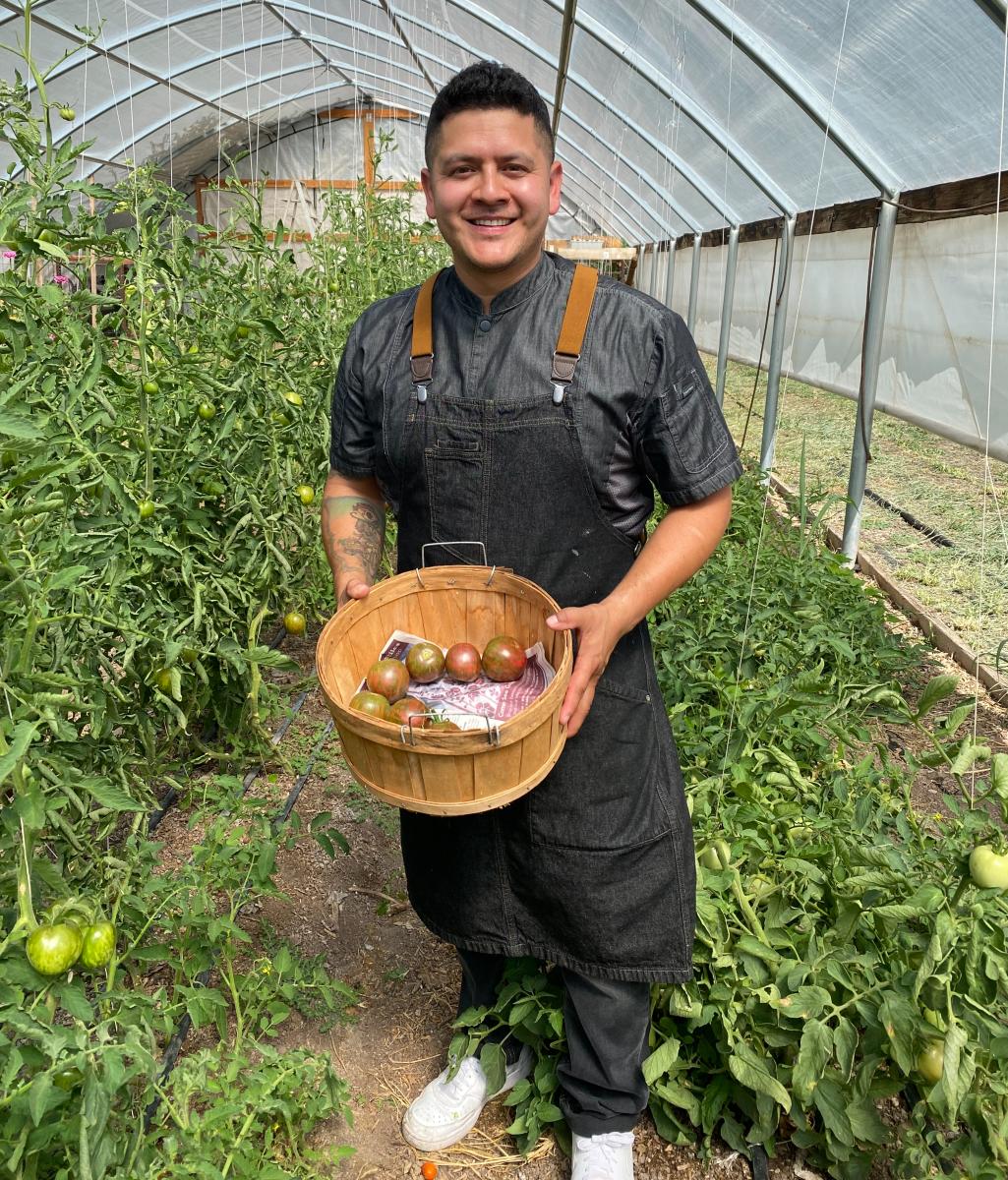 Chef David Reyes stands in a greenhouse at Bike In Coffee. He holds a basket of freshly picked tomatoes.
