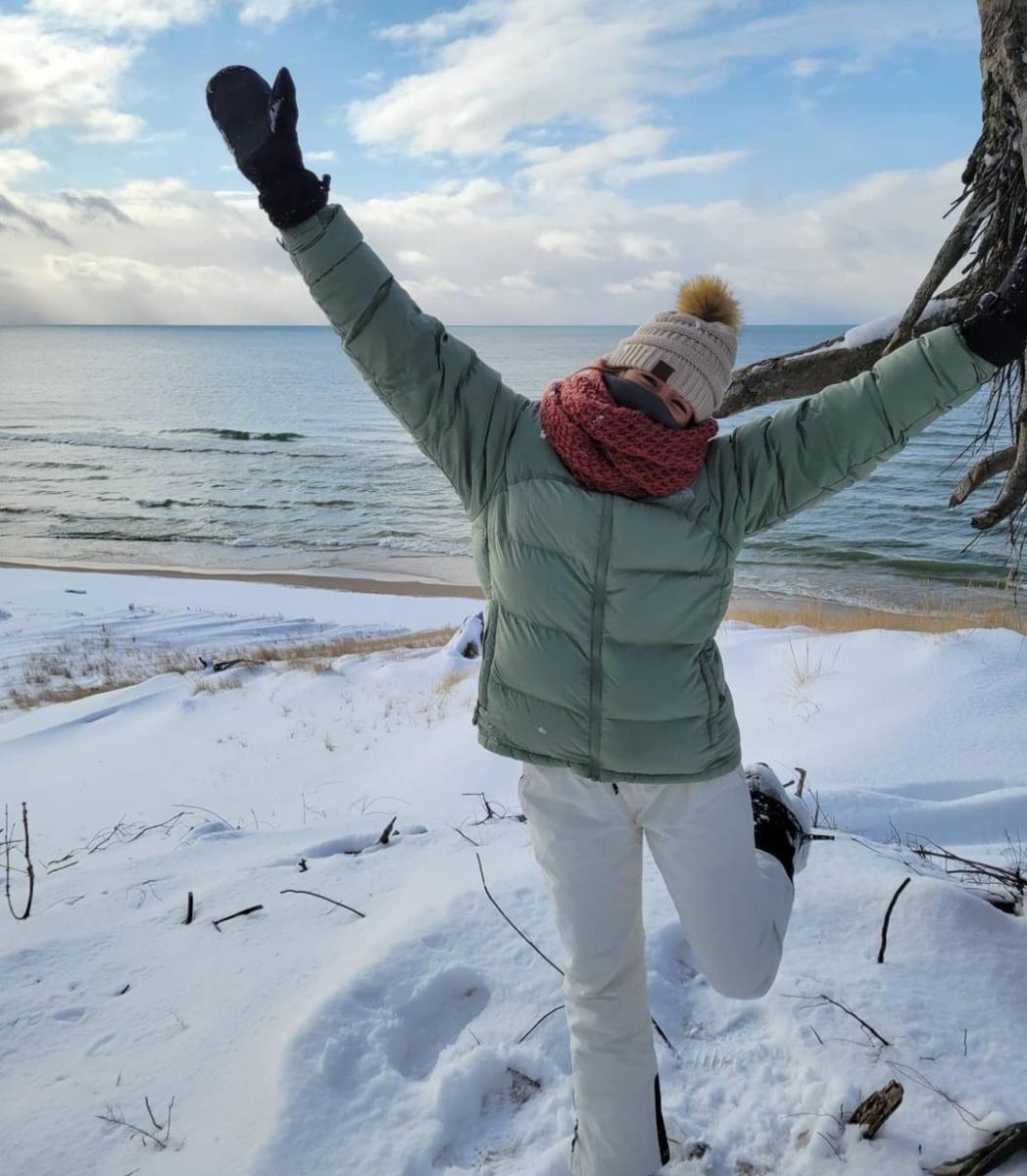 woman expresses joy while standing on snow covered hill overlooking lake michigan. she wears white snowpants, mint winter coat, burgundy scarf around her face and white knit hat.