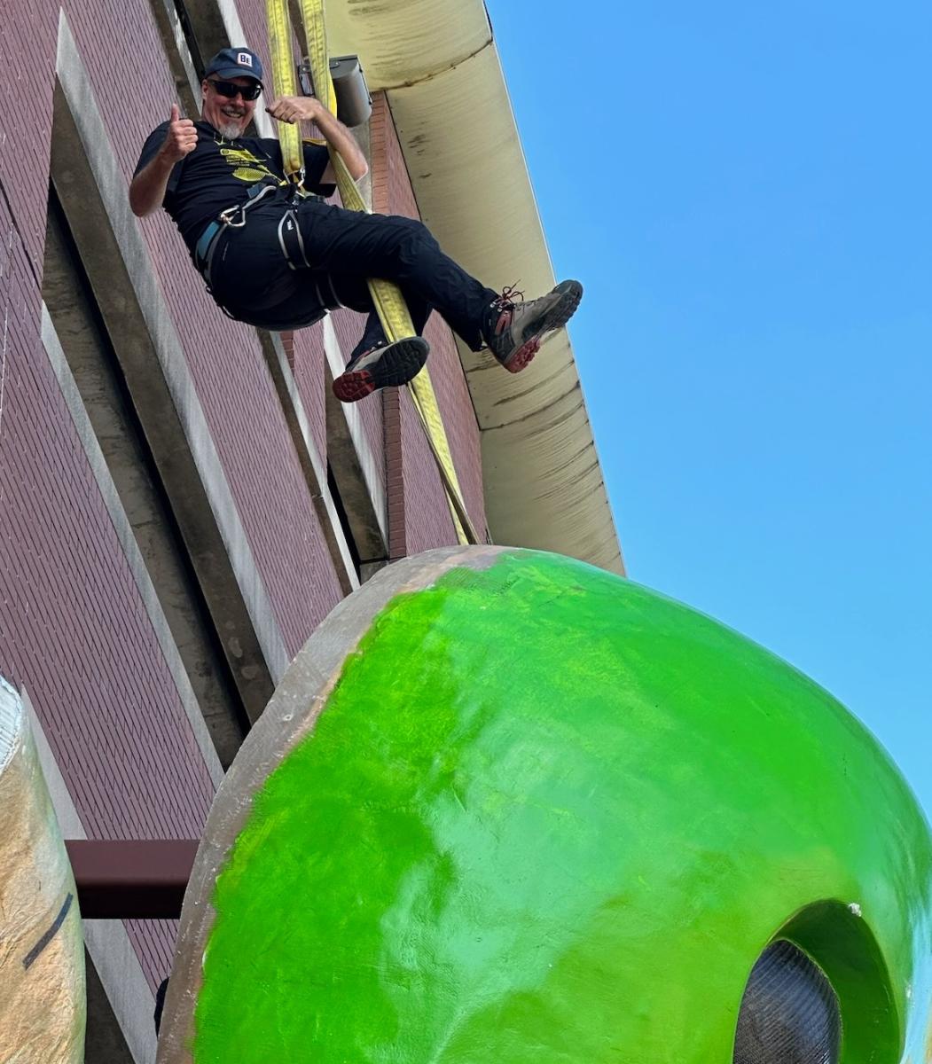Artist Marc Phelps hanging above the green head of his sculpture Clive the Alien
