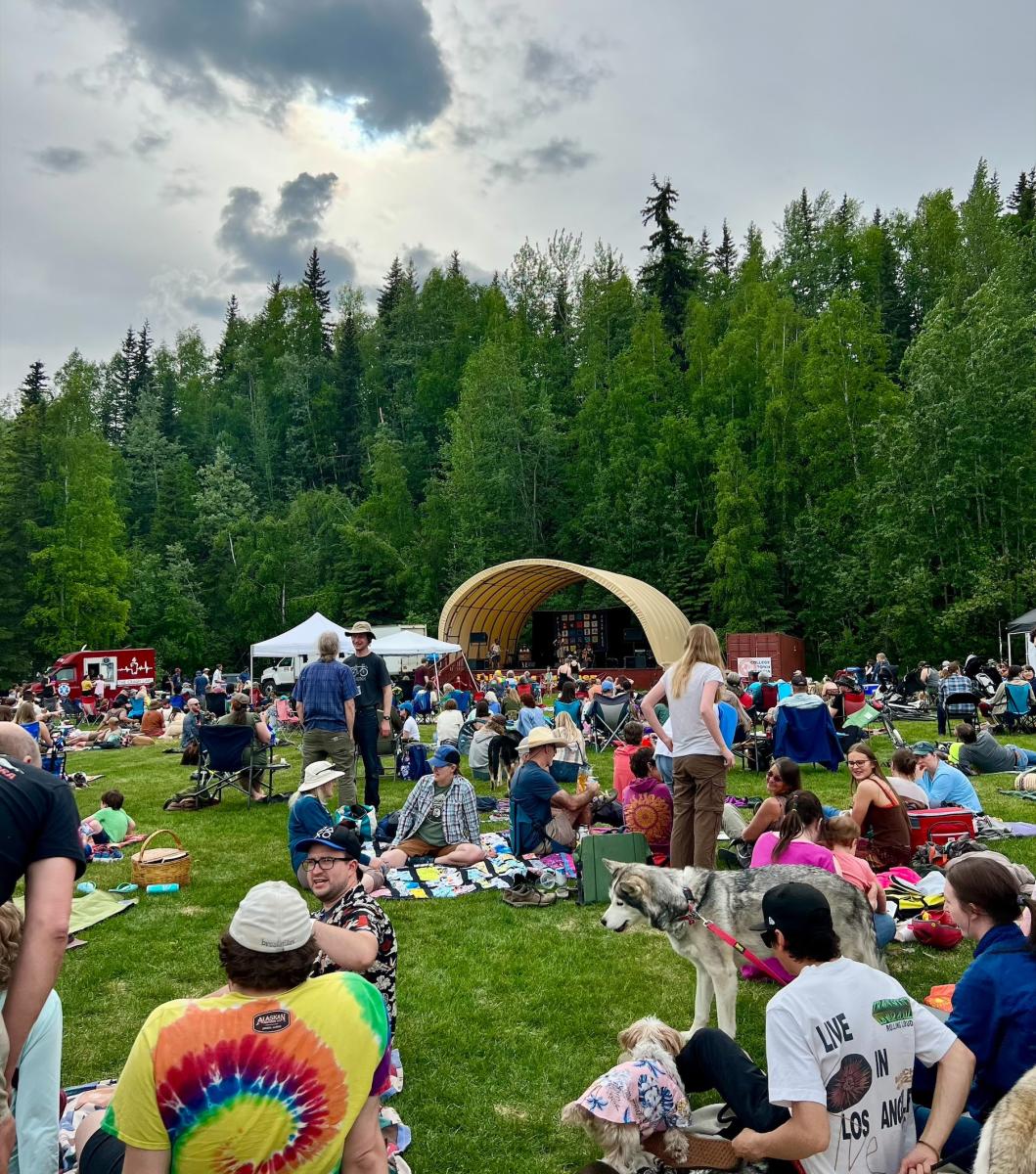 People dancing, sitting and standing at the Fairbanks Summer Folk Festival on a grassy lawn in front of an outdoor stage surrounded by green trees.