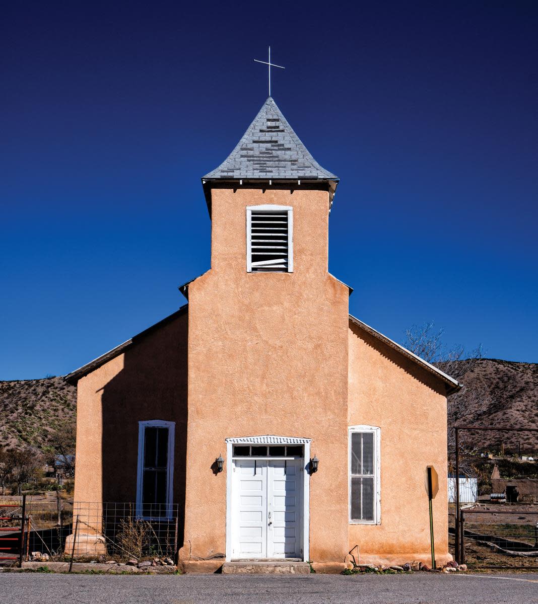 Exterior view of a historic Mission-style church with a adobe facade and a pointed steeple topped with a cross, set against a clear blue sky.