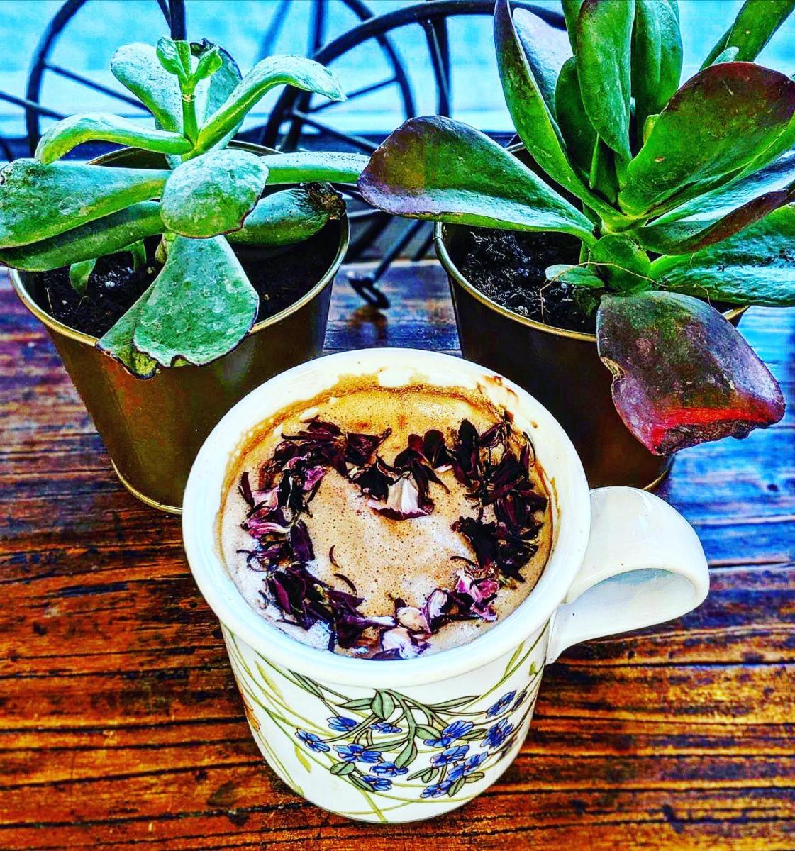 Image is a floral coffee mug with a late in it with flowers that make a heart on top and two plants next to it.