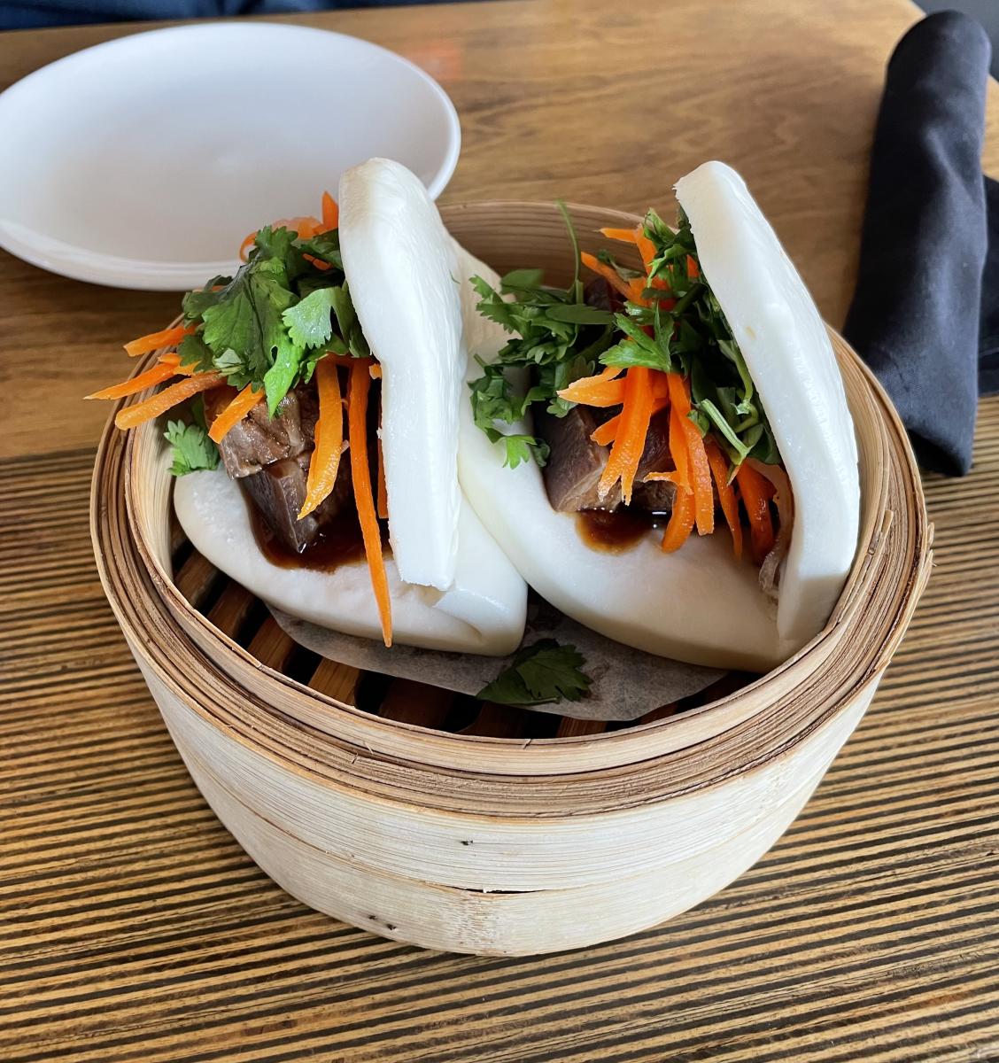 Two fluffy steamed bao buns filled with tender slices of meat, shredded carrots, fresh cilantro, and a drizzle of savory sauce, served in a bamboo steamer basket on a wooden table.