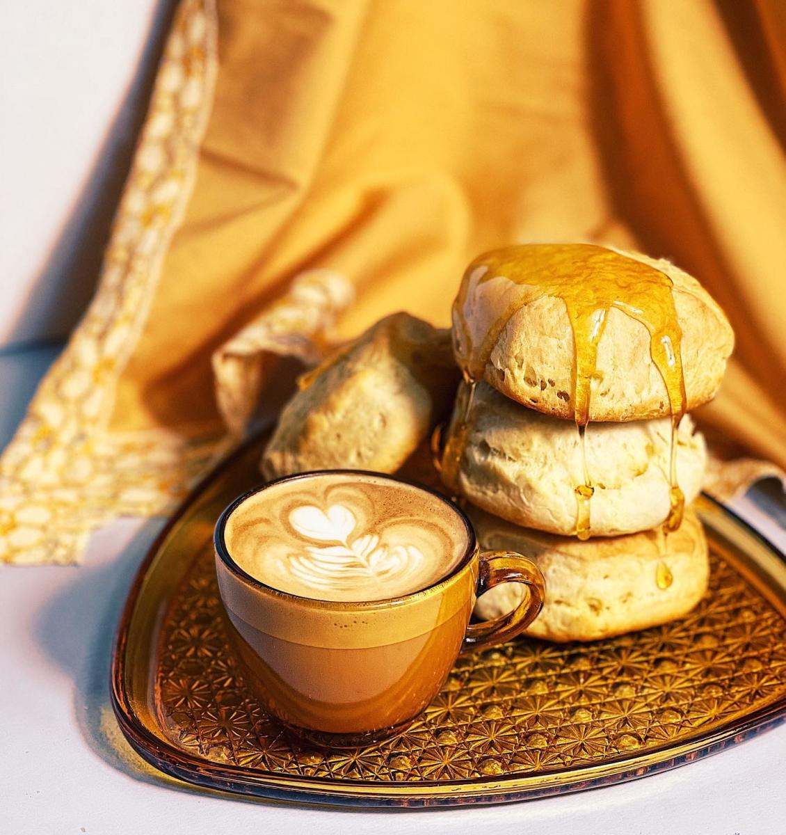 A golden-brown tray with three golden hued biscuits with honey drizzeled over it, sitting next to a brown and tan coffee mug with a late.