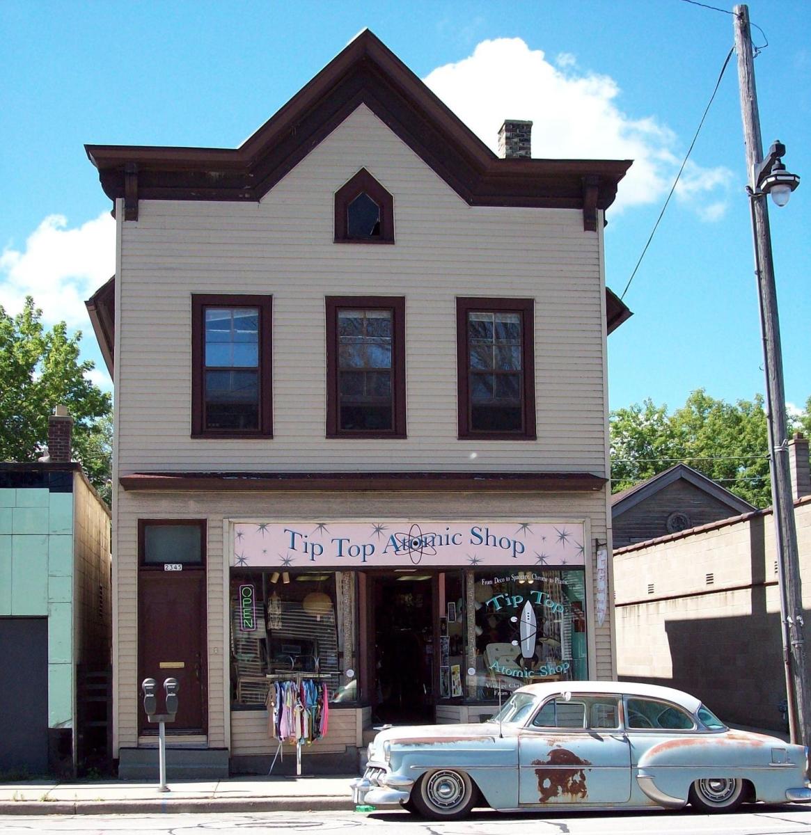 Two-story vintage shop building labeled “Tip Top Atomic Shop” with a retro sign, clothing rack outside, and a classic car parked along the street in front.