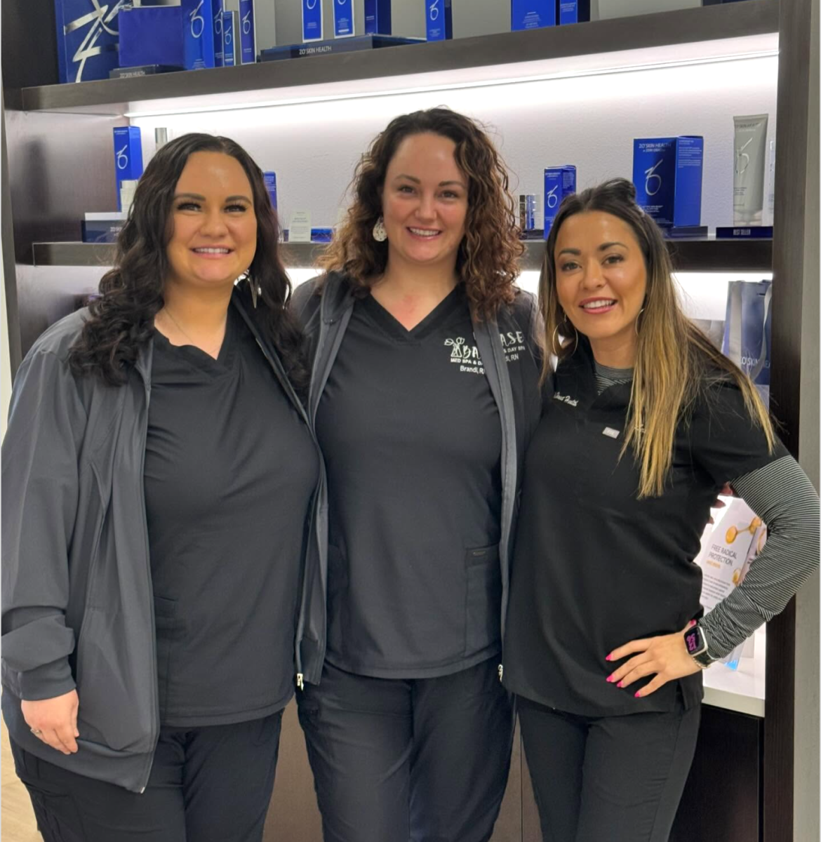 Three women wearing black medical scrubs stand together in front of a display shelf filled with blue skincare product boxes. They are smiling with their arms around each other. The woman in the middle has curly hair and wears a scrub top with embroidered text. The setting has bright lighting and a modern, professional design.