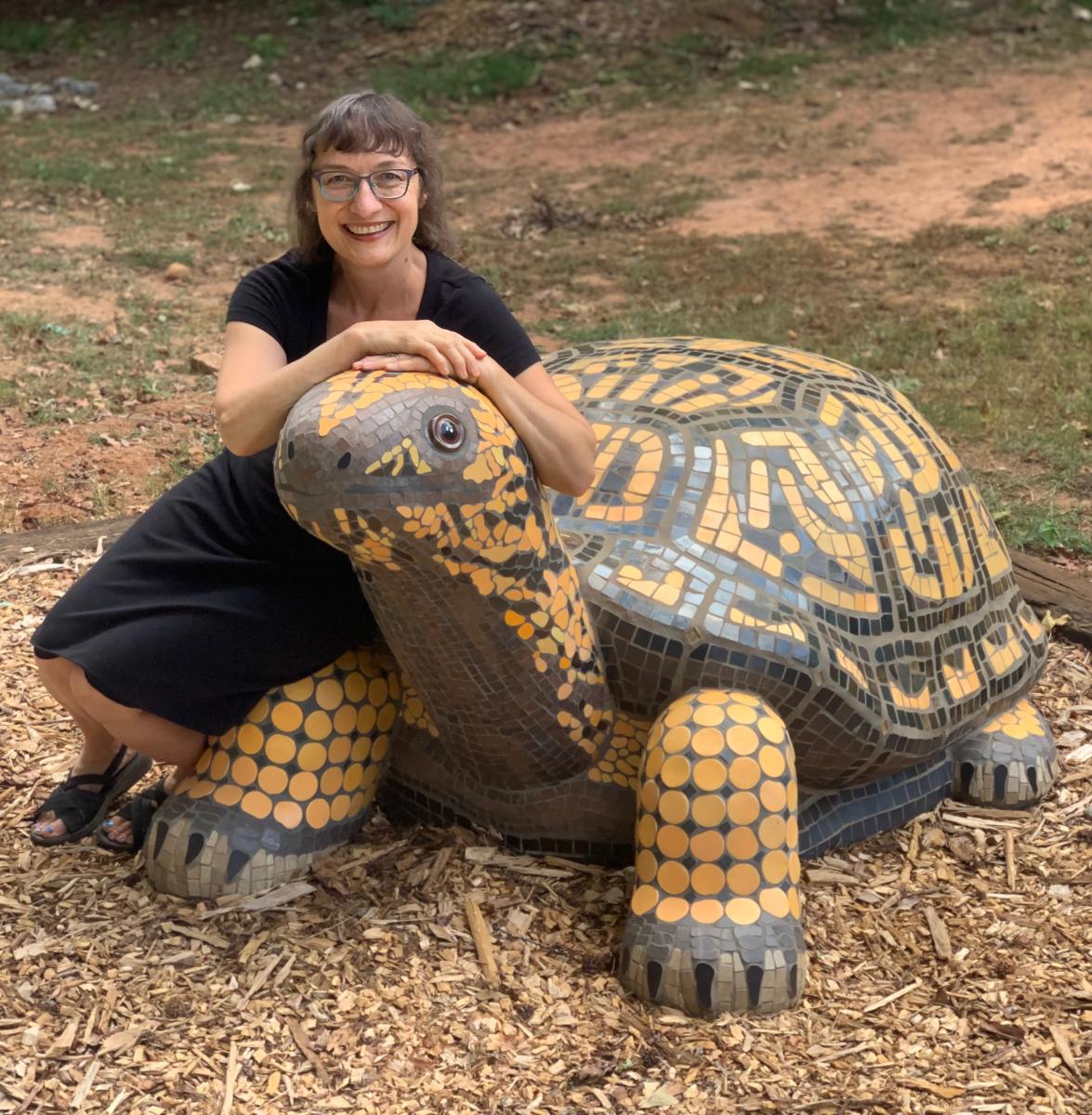 Artist Krysia Ara sits on top of a mosaic play sculpture shaped like a box turtle that she created.