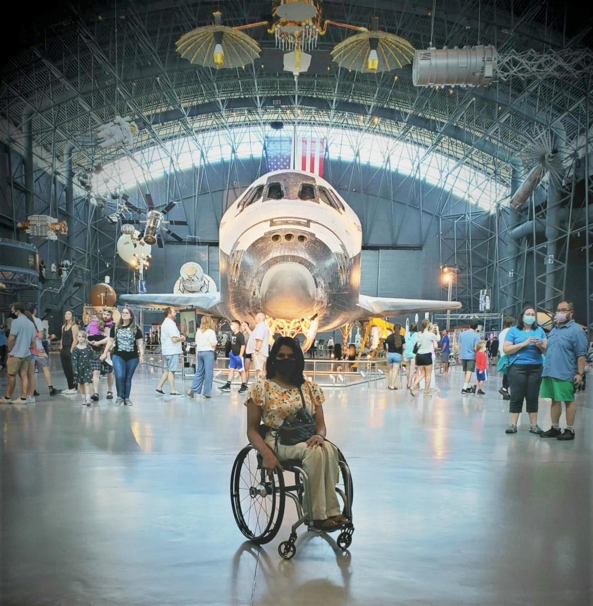 Woman in wheelchair posing in front of giant space shuttle Discovery at Air and Space Museum