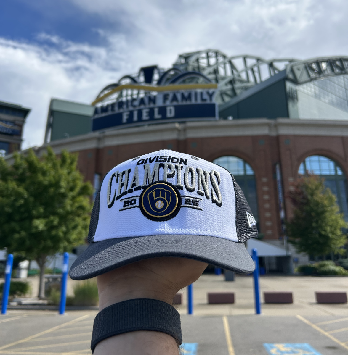 A hand holds up a Milwaukee Brewers 2025 Division Champions hat in front of American Family Field. The hat is black and white with the Brewers logo in the center. The stadium sign and retractable roof are visible in the background under a partly cloudy sky.