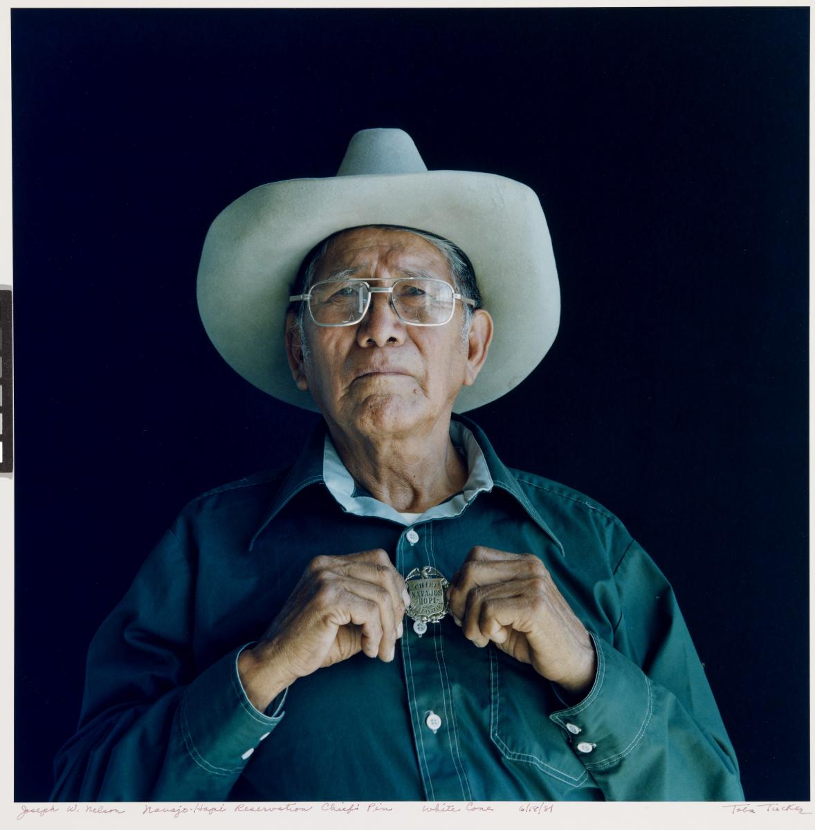 Person in a cowboy hat holding a round badge against a dark background.