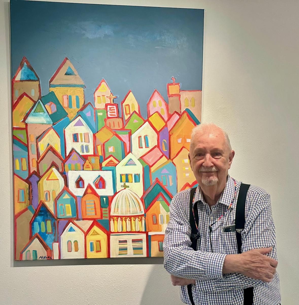 A white-haired man in a white-checkered shirt (Jim N. Hill) stands in front of one of his paintings displayed in TWAC's exhibit space. The painting depicts a colorful skyline of overlapping houses and churches of various faiths.