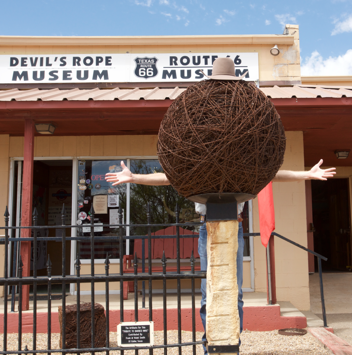 A person playfully poses behind a giant ball of barbed wire at the Devil’s Rope Museum and Route 66 Museum in McLean, Texas, with arms stretched wide as if they’re part of the sculpture. The museum’s tan exterior and classic signage hint at the quirky charm and Western heritage tucked inside this Route 66 roadside stop.
