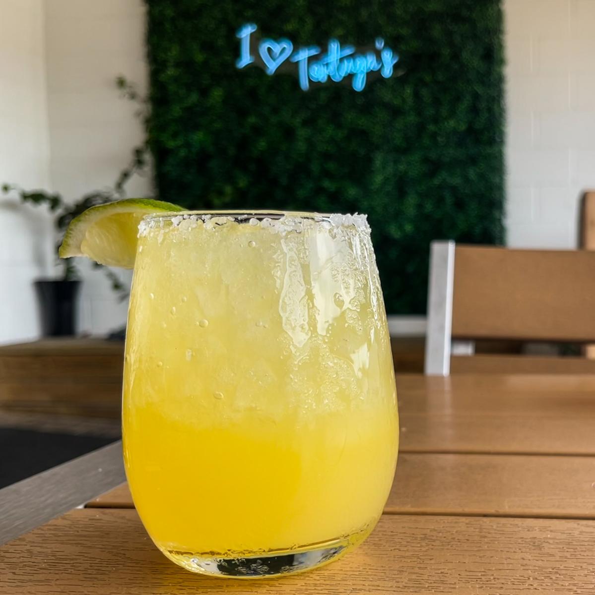 A yellow margarita in a stemless glass sits on a table. In the background is a greenery wall with a neon sign that says "I <3 Tortuga's"