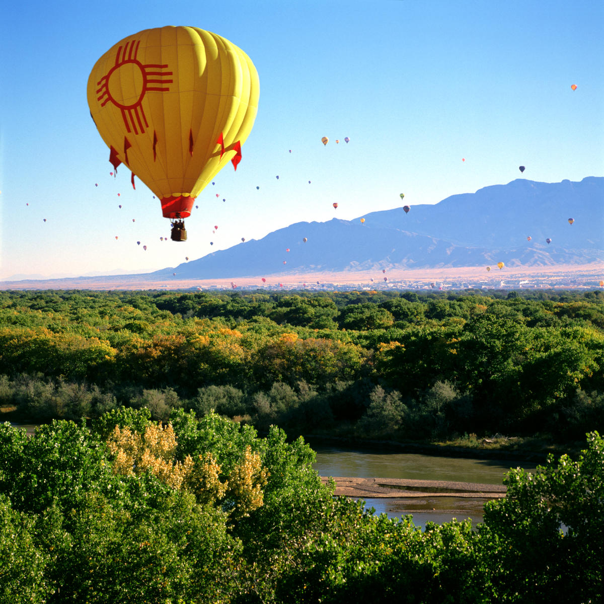 A hot air balloon with a Zia symbol floats over the Rio Grande with the Sandia Mountains in the background.