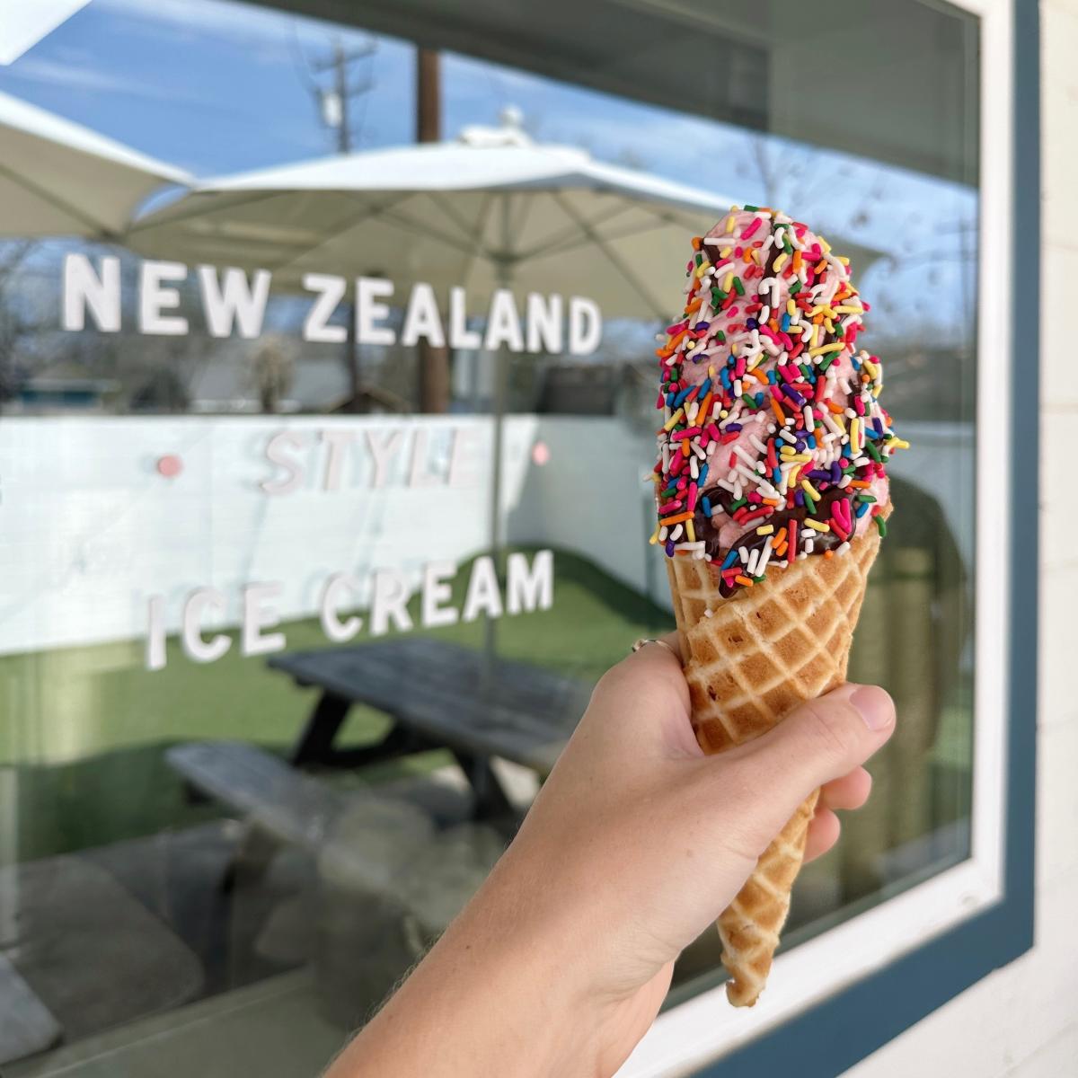 Image of a hand holding a ice cream cone with sprinkles out front of Zed's Ice Cream.