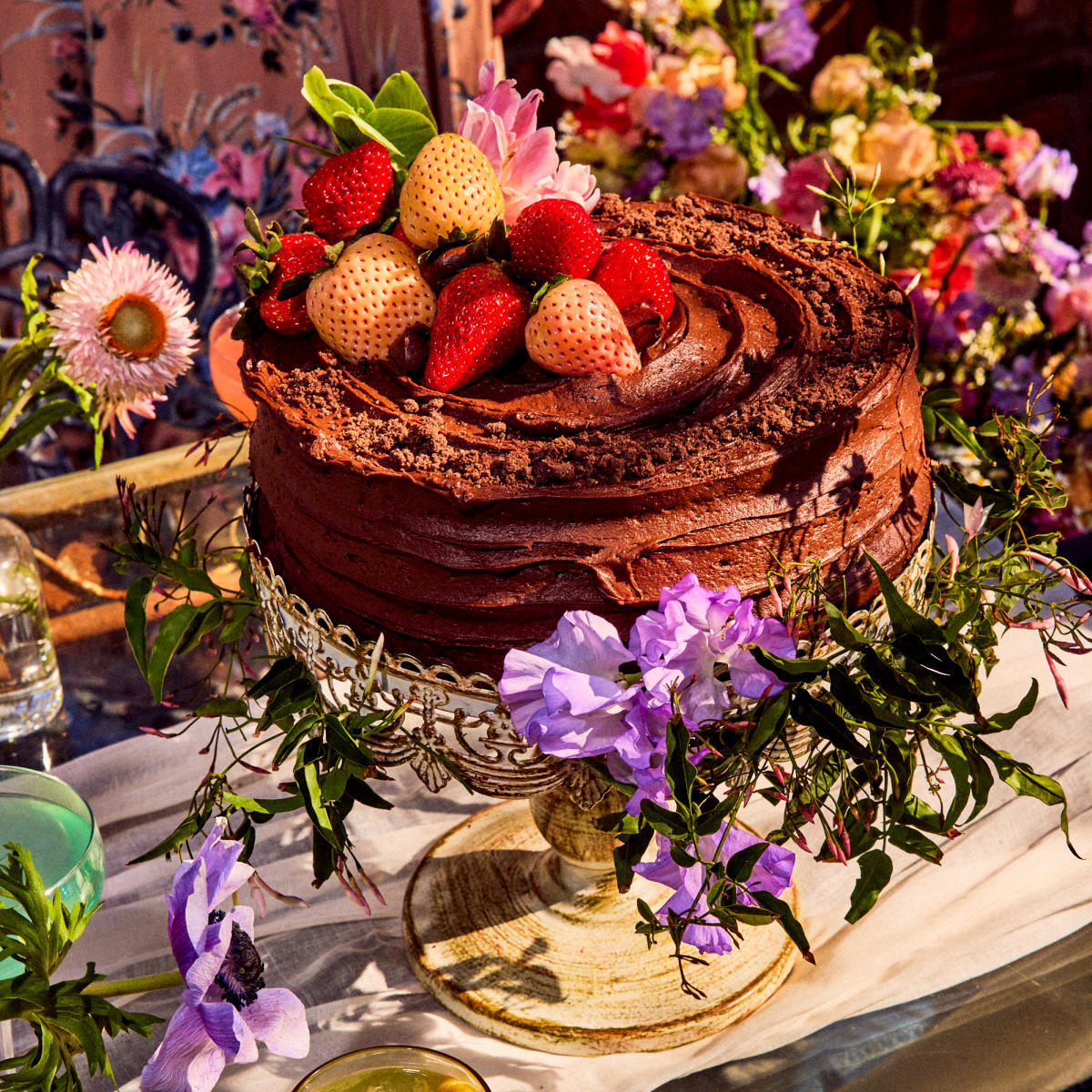 Image of a chocolate cake sitting on a pedestal adorned with colorful fruit and flowers.
