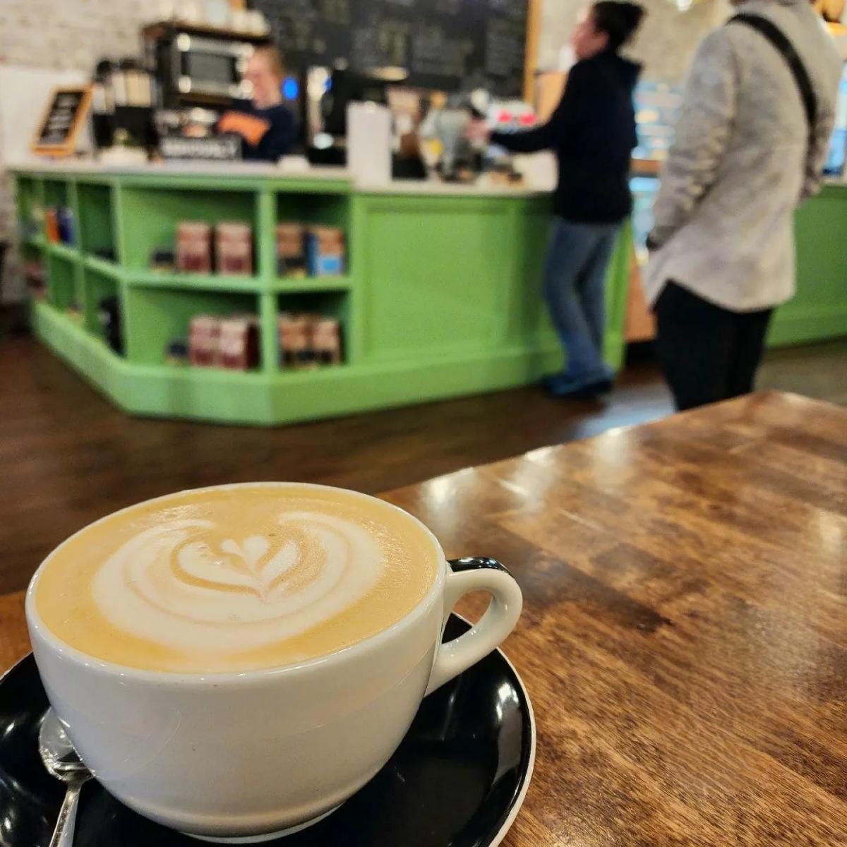 Image is of late with foam in a leaf design, in a white mug with people and the counter in the background.