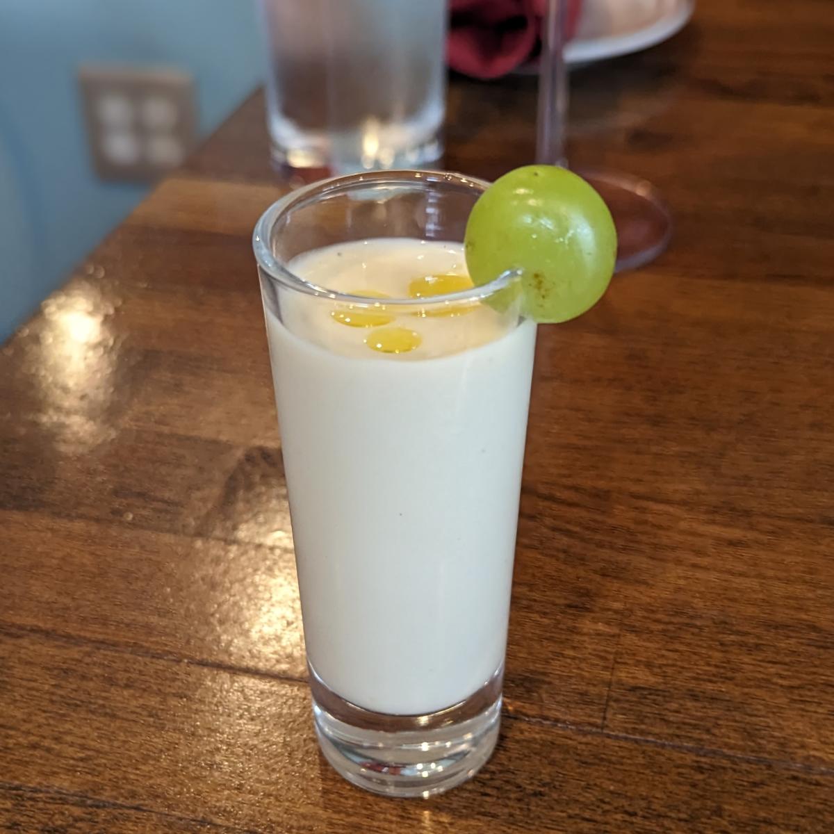 The image is of a Spanish cucumber smoothie in a shot glass with a grape on the rim.
