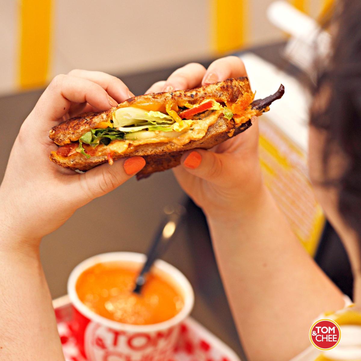 Image is of a person holding a grilled cheese sandwich in her hand and a cup of tomato soup in front of her.