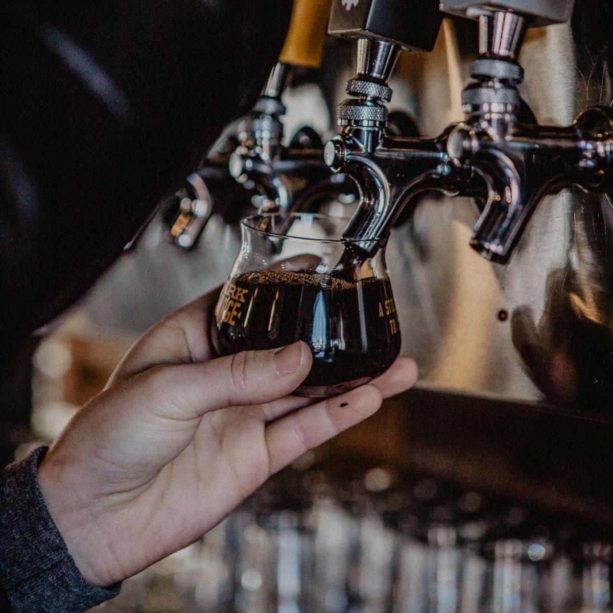 A hand holding a Braxton taster glass up to the craft beer taps for a pour of Dark Charge Imperial Stout in Covington, Ky.