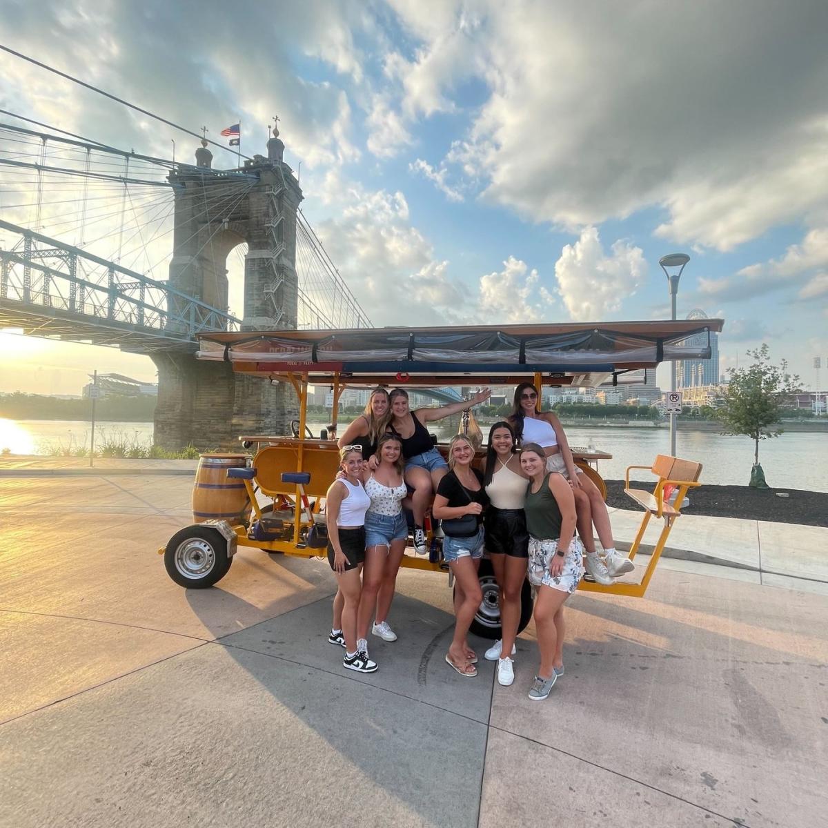 A group of young women standing and smiling in front of the Covington Pedal Wagon at the Roebling Suspension bridge
