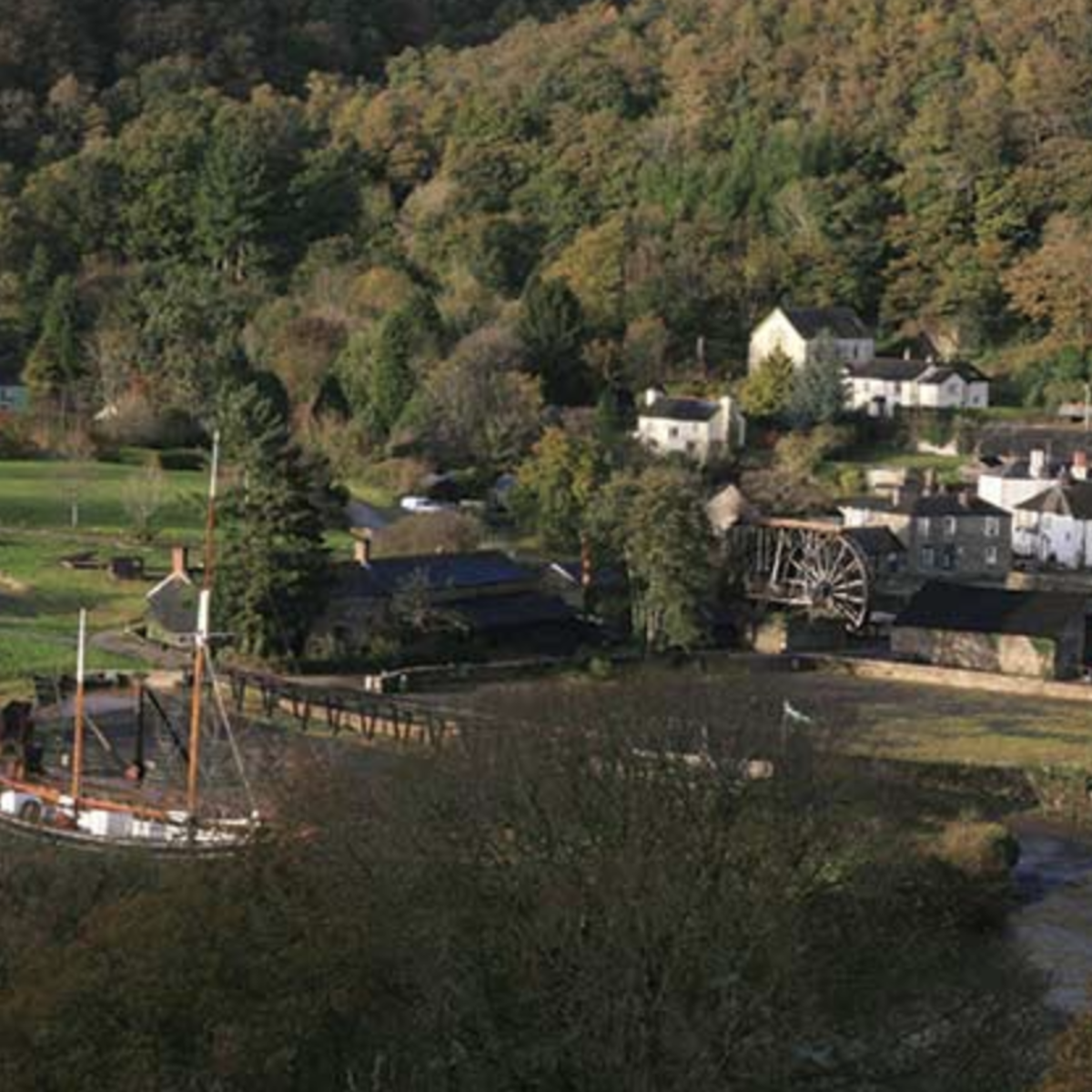 A scenic village nestled in a lush, forested valley. Features quaint cottages, a waterwheel, open green fields, and a docked ship, evoking tranquillity.