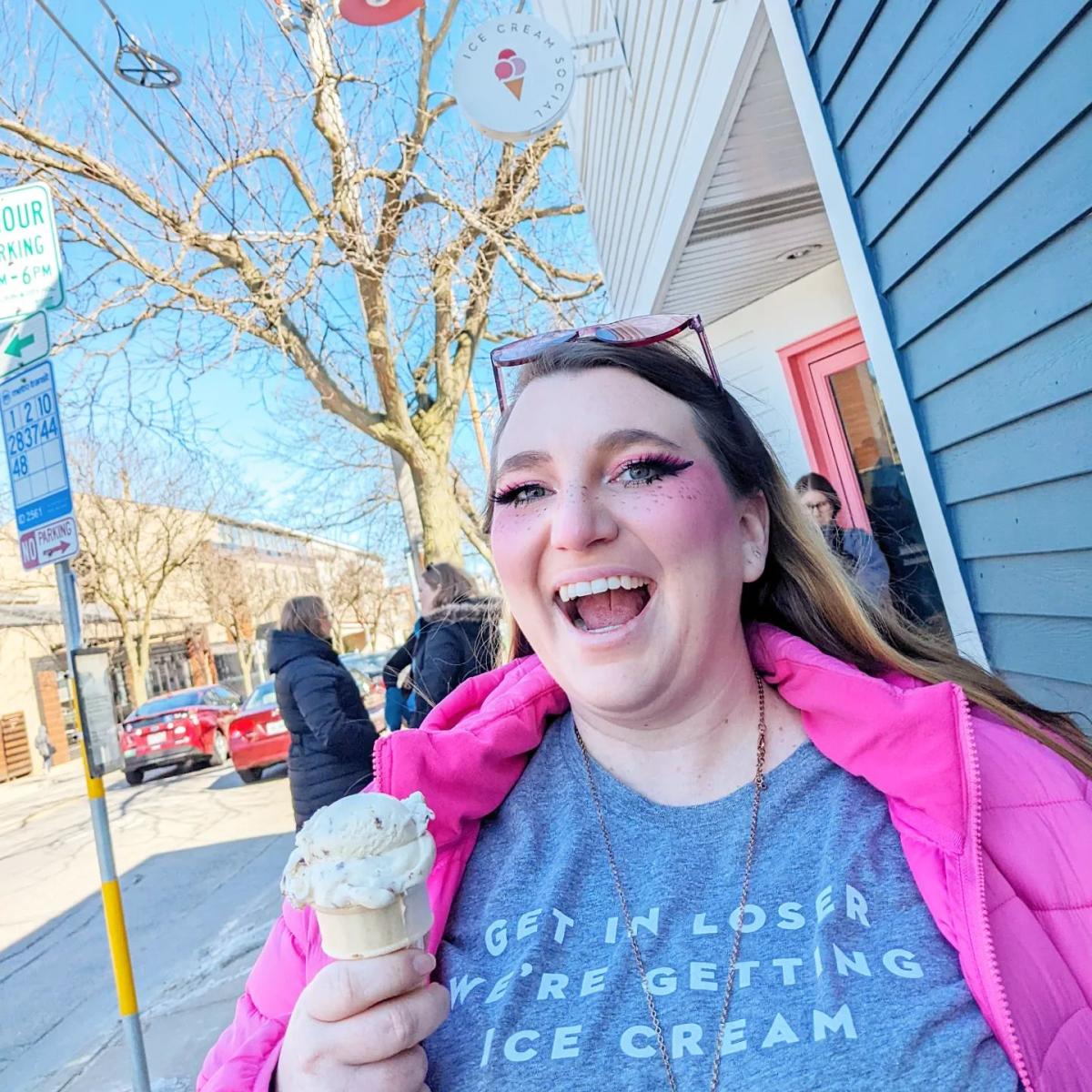A white woman with bright makeup on and sunglasses on her head smiles widely while carrying an ice cream cone outside of Ice Cream Social