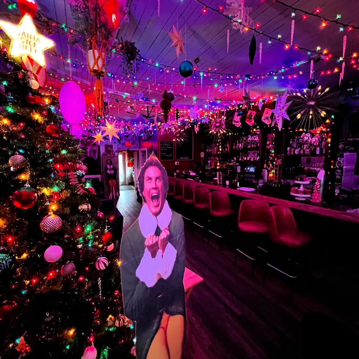 A bar decorated with colorful holiday lights that cover the ceiling in long strands along with stars, ornaments, and icicle shaped lights. A large Christmas tree filled with ornaments and glowing bulbs stands on the left. In front of the tree is a life-size cutout of Buddy the Elf with an excited expression. Red bar stools line the counter on the right where more holiday decorations surround bottles and shelves. The room glows with pink and purple lighting that adds to the festive atmosphere.
