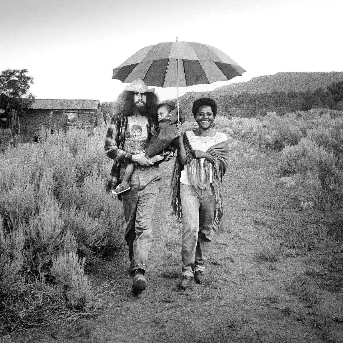 Black and white photograph of three people walking along a dirty path beneath a striped umbrella, including a man carrying a small child and a smiling woman.