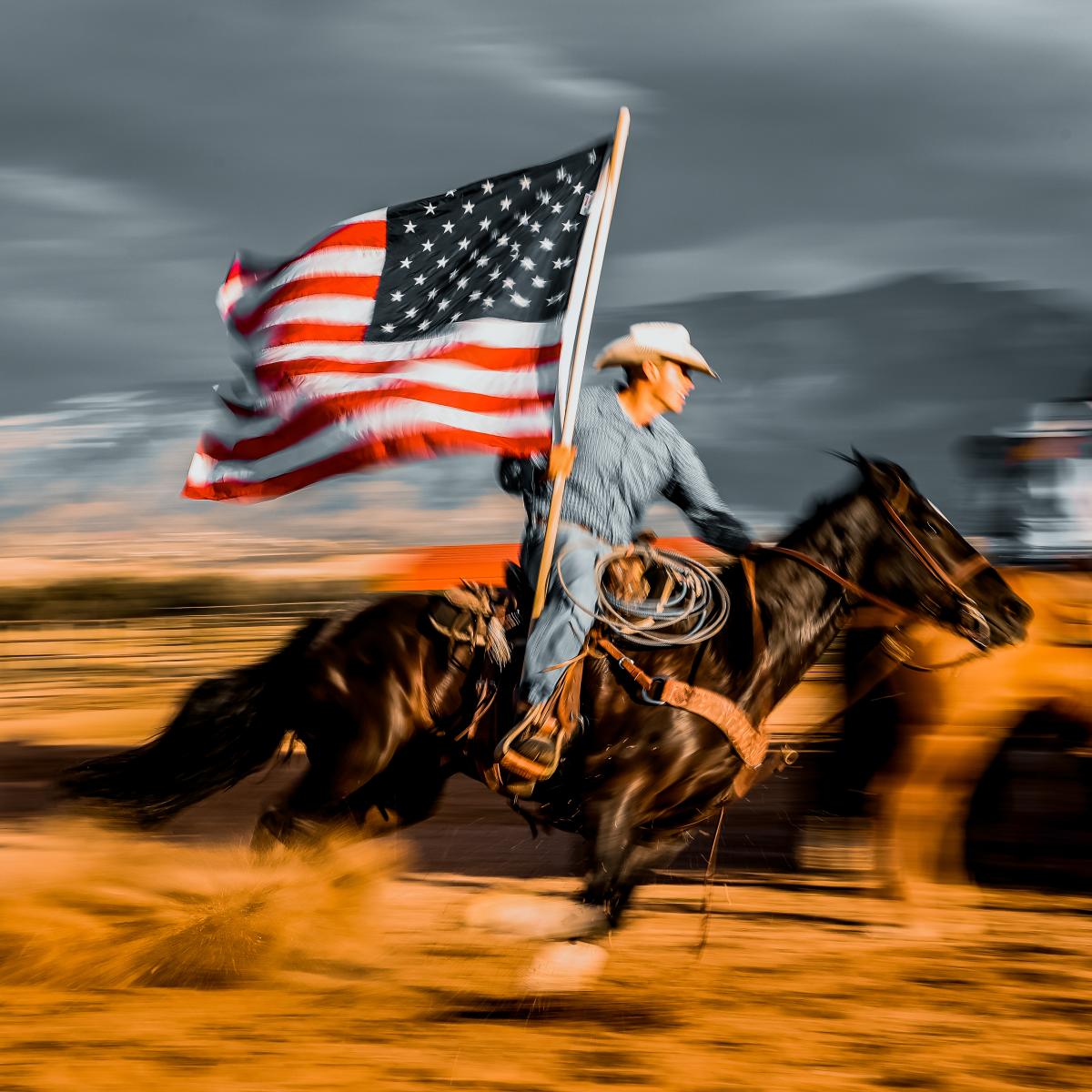 A rodeo rider carrying an American flag at full gallop across a dusty area.