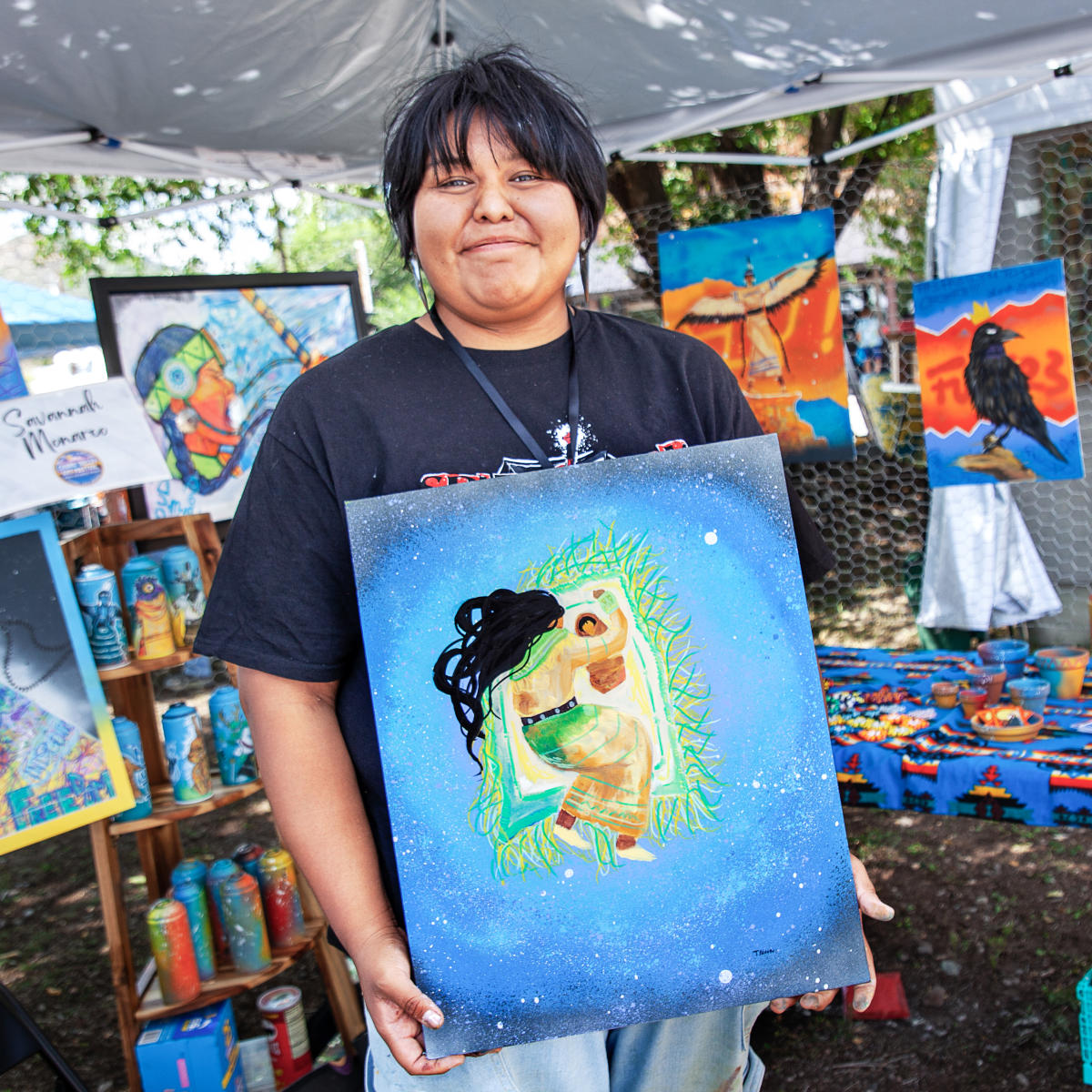 Artist at an outdoor booth holding a colorful painting, with additional artwork and supplies displayed behind.