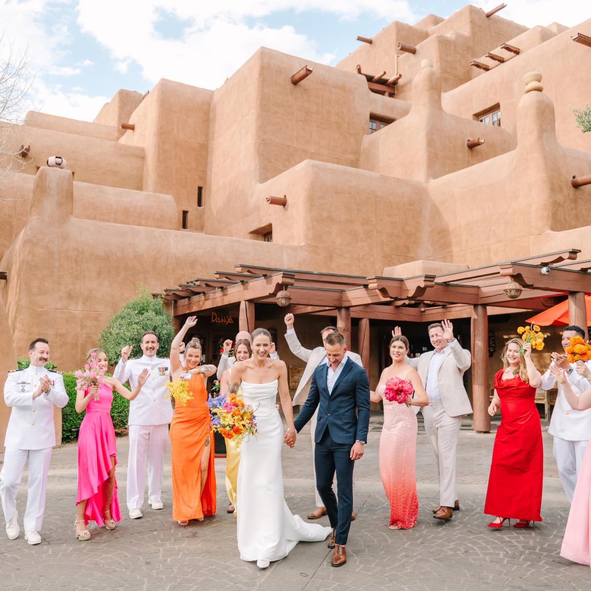 Bride and groom happily walk with a cheering wedding party in colorful attire. They’re set against a backdrop of adobe-style buildings under a blue sky.