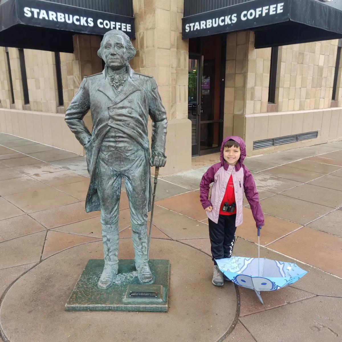 A child in a purple raincoat stands smiling next to a bronze statue of a historical figure outside a Starbucks Coffee. The child holds an open umbrella with a playful design. The scene is lively and casual.