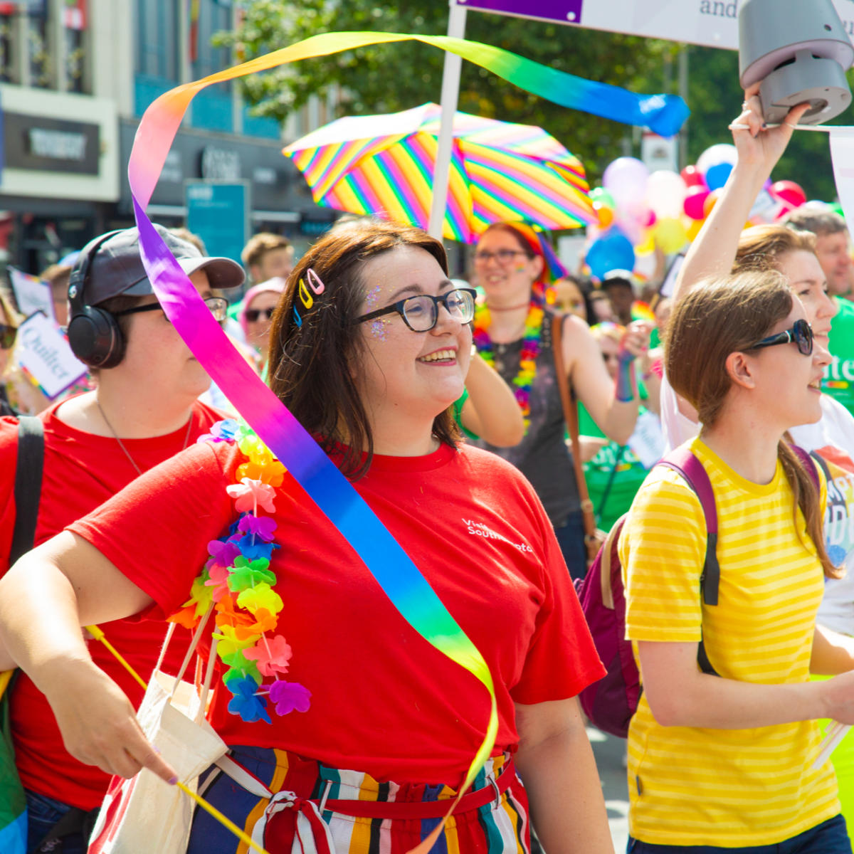 Woman smiling waving rainbow ribbon in Southampton Pride march through Above Bar Street