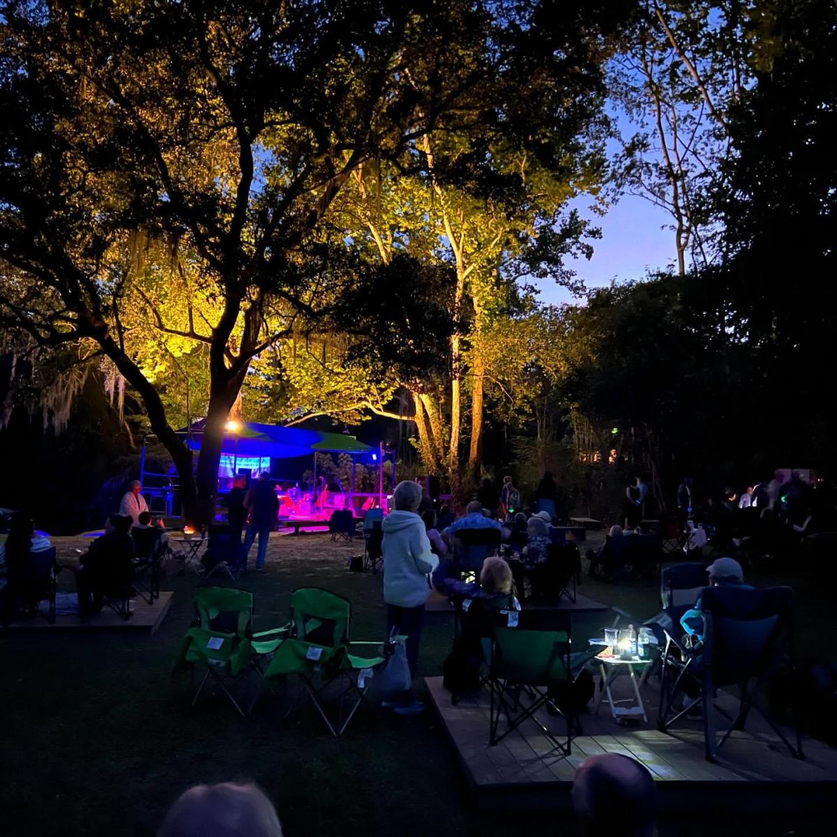 Crowd seated in lawn chairs beneath oak trees at Columbia Street Landing during a Sunset at the Landing concert, with colorful stage lights glowing and the Bogue Falaya River just behind the stage.