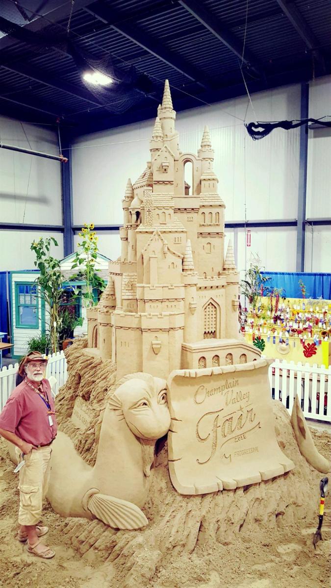 A man in khakis and a red shirt stands next to a sand sculpture double his height. The sculpture is a castle with a monster in the moat and a scroll that says "Champlain Valley Fair"