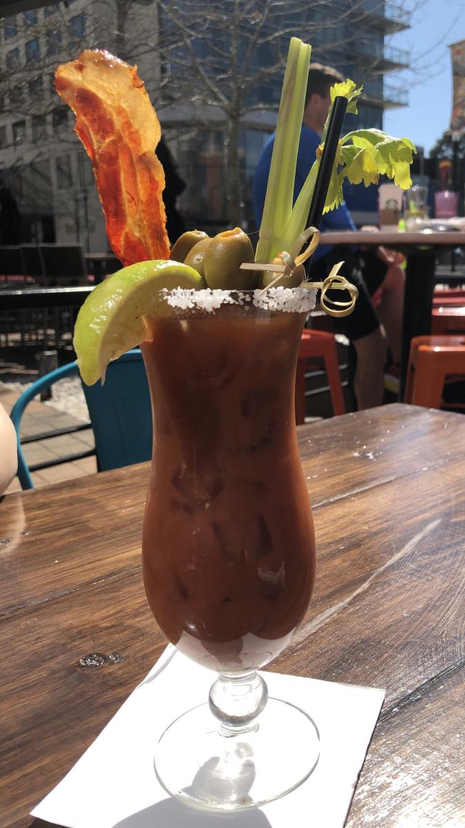 On a sunny patio, a tall drink stands on a sturdy wooden table. The drink is dark orange or brown in color, salt-rimmed and piled high with celery stalks, a slice of bacon, a lime wedge, and a stick of olives balanced across the lip. In the background are two men sharing drinks at another table.