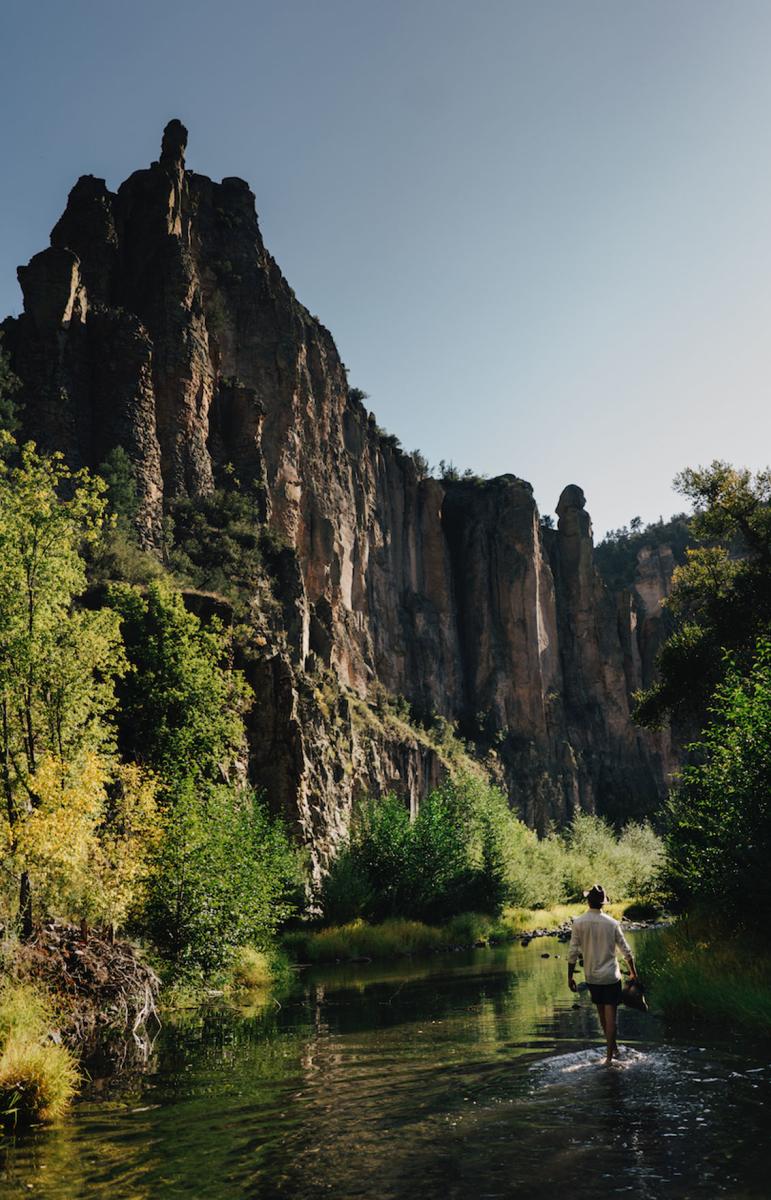 A person walks through a shallow river with tall, rugged cliffs on either side, under a clear sky.
