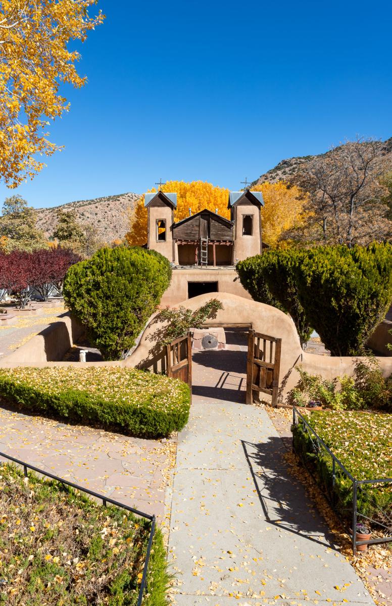 A historic adobe church with tall towers sits behind a courtyard with adobe walls and a gate. There are trees with fall foliage and bushes and shrubs surrounding the church.