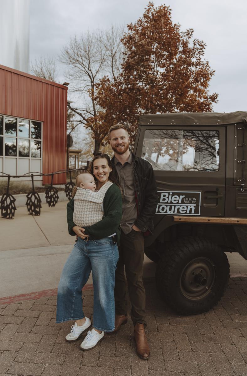 A man standing with a woman who is wearing a baby in front of a pinzgauer that says "Bier Touren" on the side.