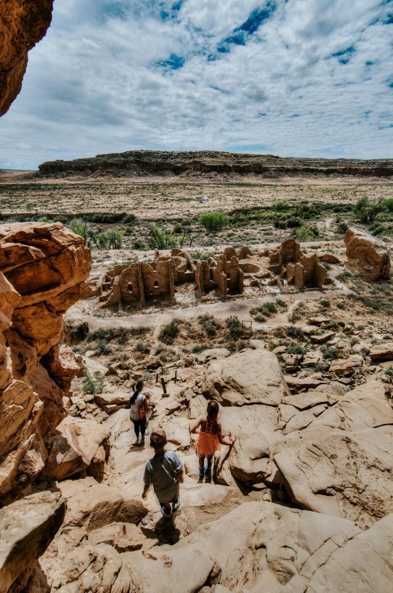 A group of people navigate a rocky cliffside looking over a desert landscape with a large mesa in the distance