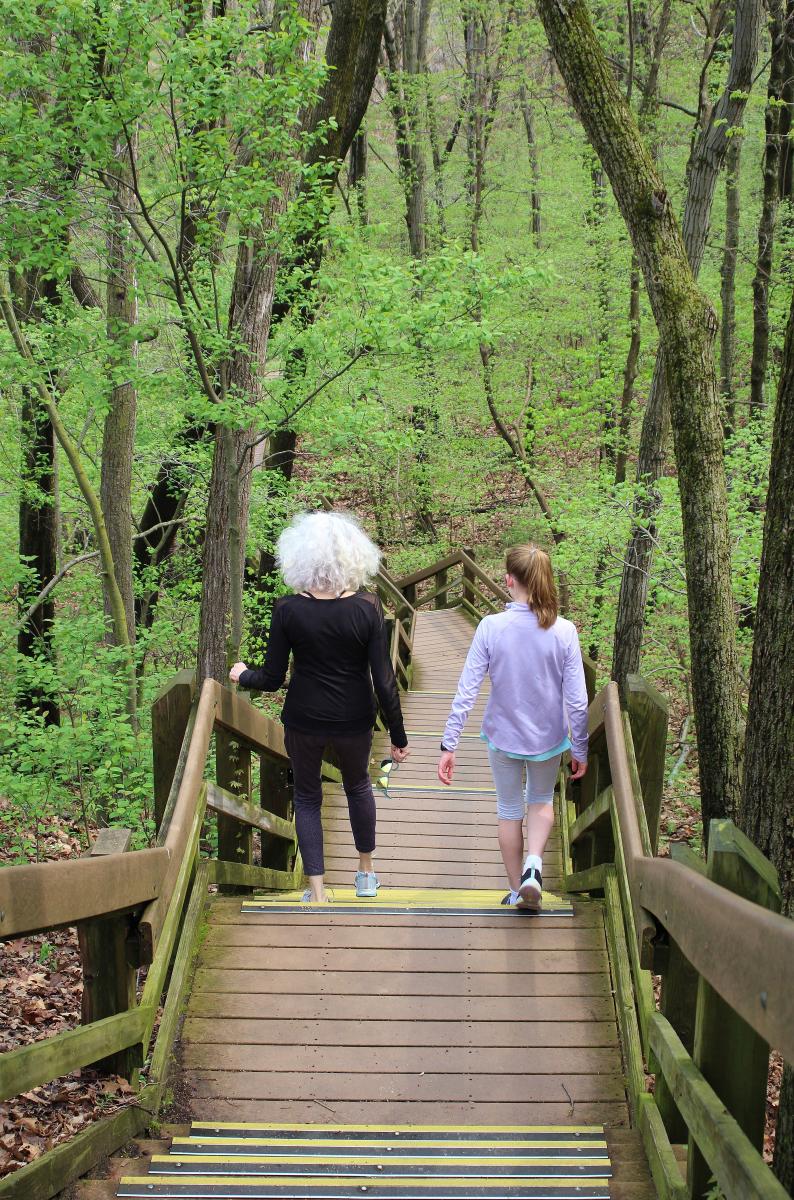 A woman with gray curly hard and a young teen descend down a wooden staircase while surrounded by woods