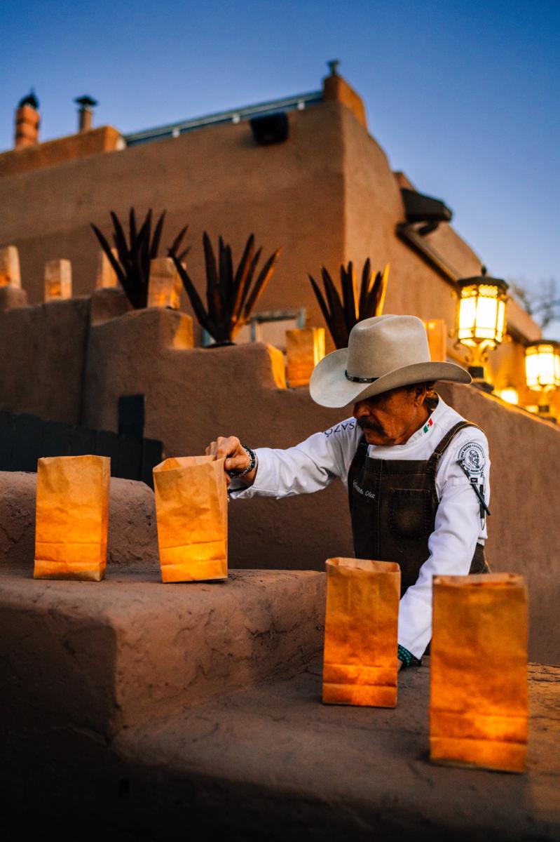 Fernando olea, in chef attire and a cowboy hat adjusting a luminaria or traditional paper bag with a flame inside that is sitting on an adobe wall with a tall adobe building behind it.